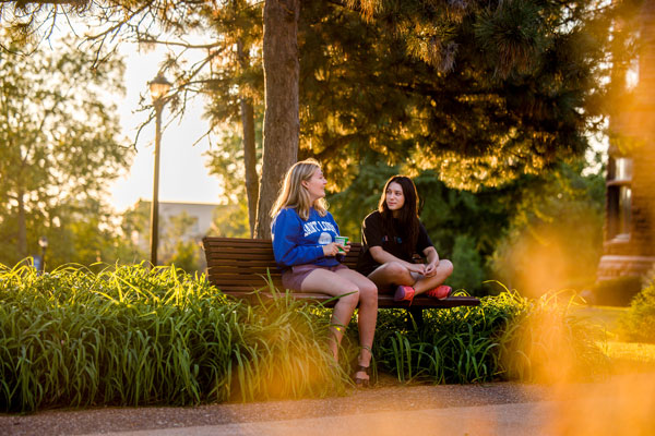 Two students seated on a bench talking, with trees and plants around them