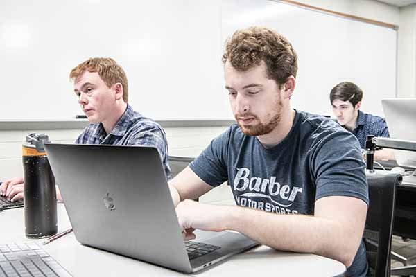 Two male students take notes on their laptops at a desk during class.