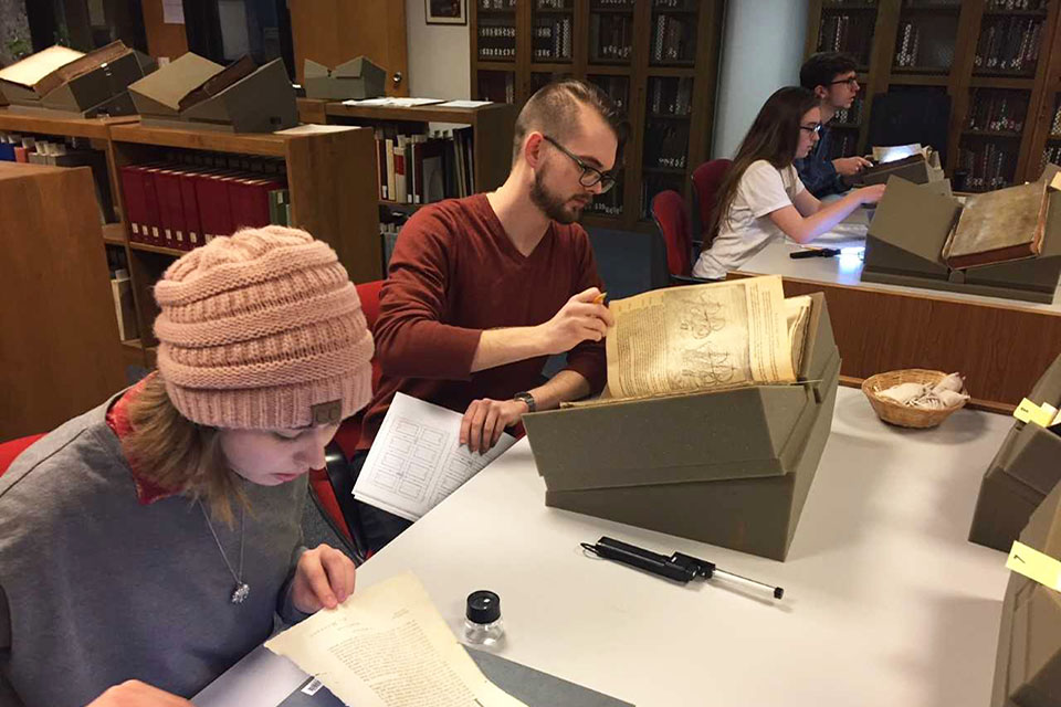 Students sit at tables in a library, examining old texts.