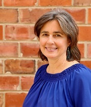 Leah Sweetman, headshot, wears a blue blouse and stands in front of a red brick wall, smiling.