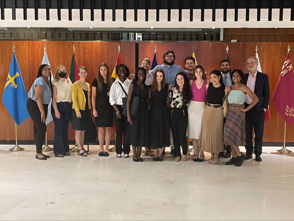 Students stand before a wall in a government building with flags in the background.