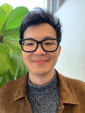 Young man with dark hair in glasses standing in front of a large window and plant.