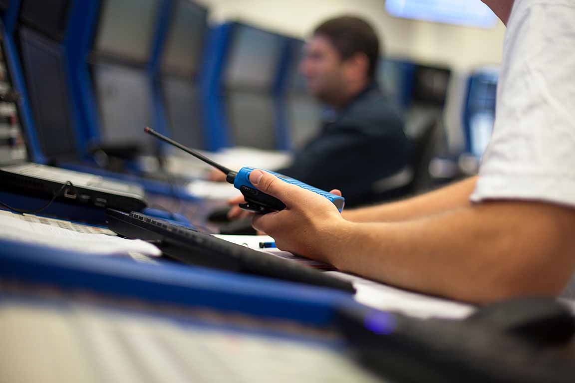 A hand holding a walkee talkee at a desk with a computer with a man in the background