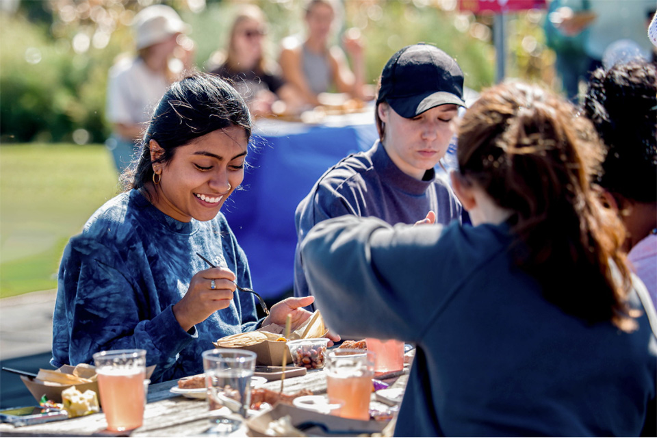 SLU students sit at a picnic table filled with cups and cardboard trays filled with food on a sunny day.