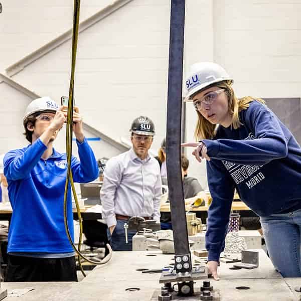 A female student wearing a hard hat points to a bend in a beam in a lab. Another male student, also wearing a hard hat, takes photos of the beam on a cell phone. A male instructor can be seen behind them watching them work.